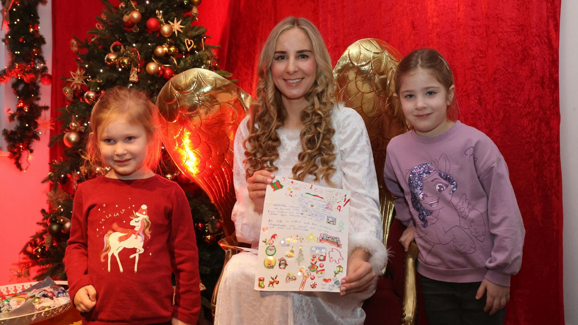 Zwei Kinder stehen links und rechts neben einer Christkinddarstellerin, die auf einem goldenen Stuhl sitzt und Engelsflügel trägt.
