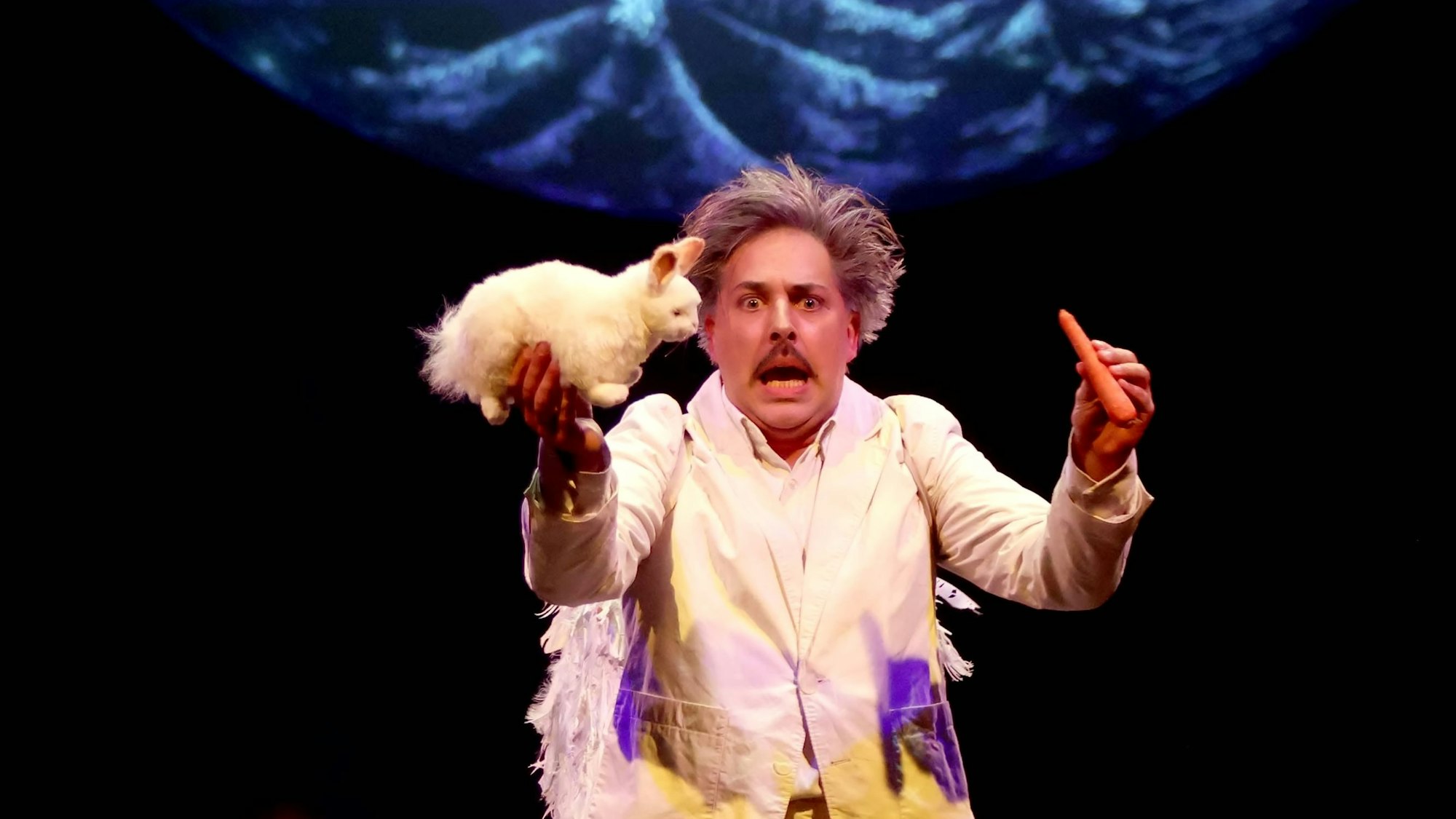 Ein Clown im weißen Outfit hält ein Stoffkaninchen und eine Möhre.