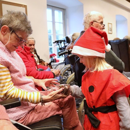 Ein Kind mit roter Nikolausmütze übergibt einer Seniorin ein kleines Geschenk.