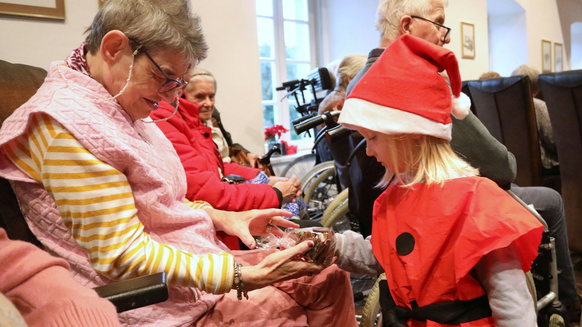 Ein Kind mit roter Nikolausmütze übergibt einer Seniorin ein kleines Geschenk.