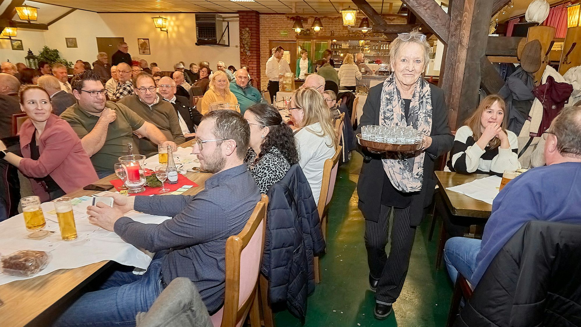 Eine Kellnerin trägt ein Tablett voller Schnapsgläser durch den Saal einer voll besetzten Gaststätte.