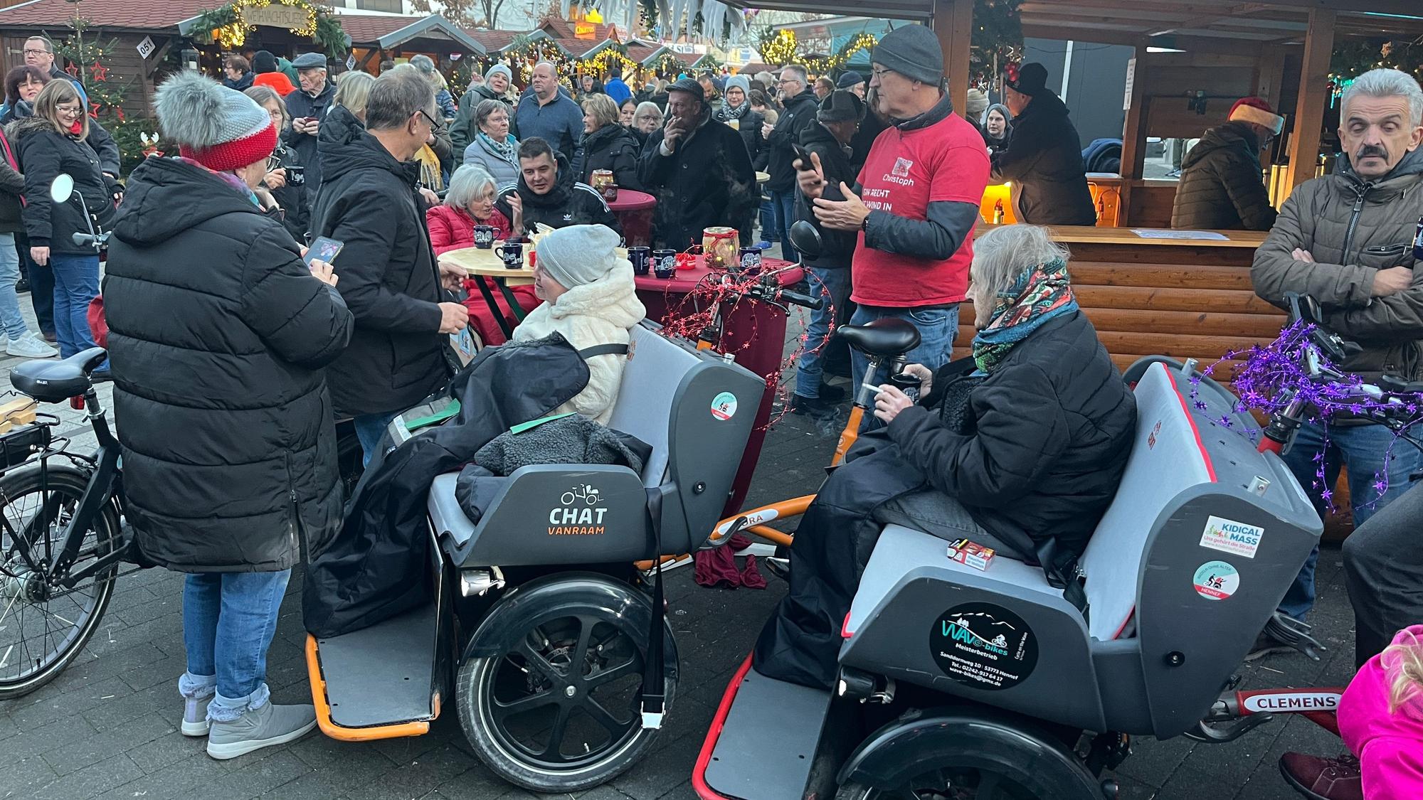 Mehrere Rikschas mit älteren Menschen stehen vor einer Bude auf einem Weihnachtsmarkt.