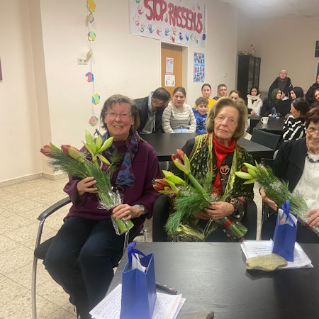 Drei ältere Frauen sitzen vor einem Tisch und halten Blumensträuße.