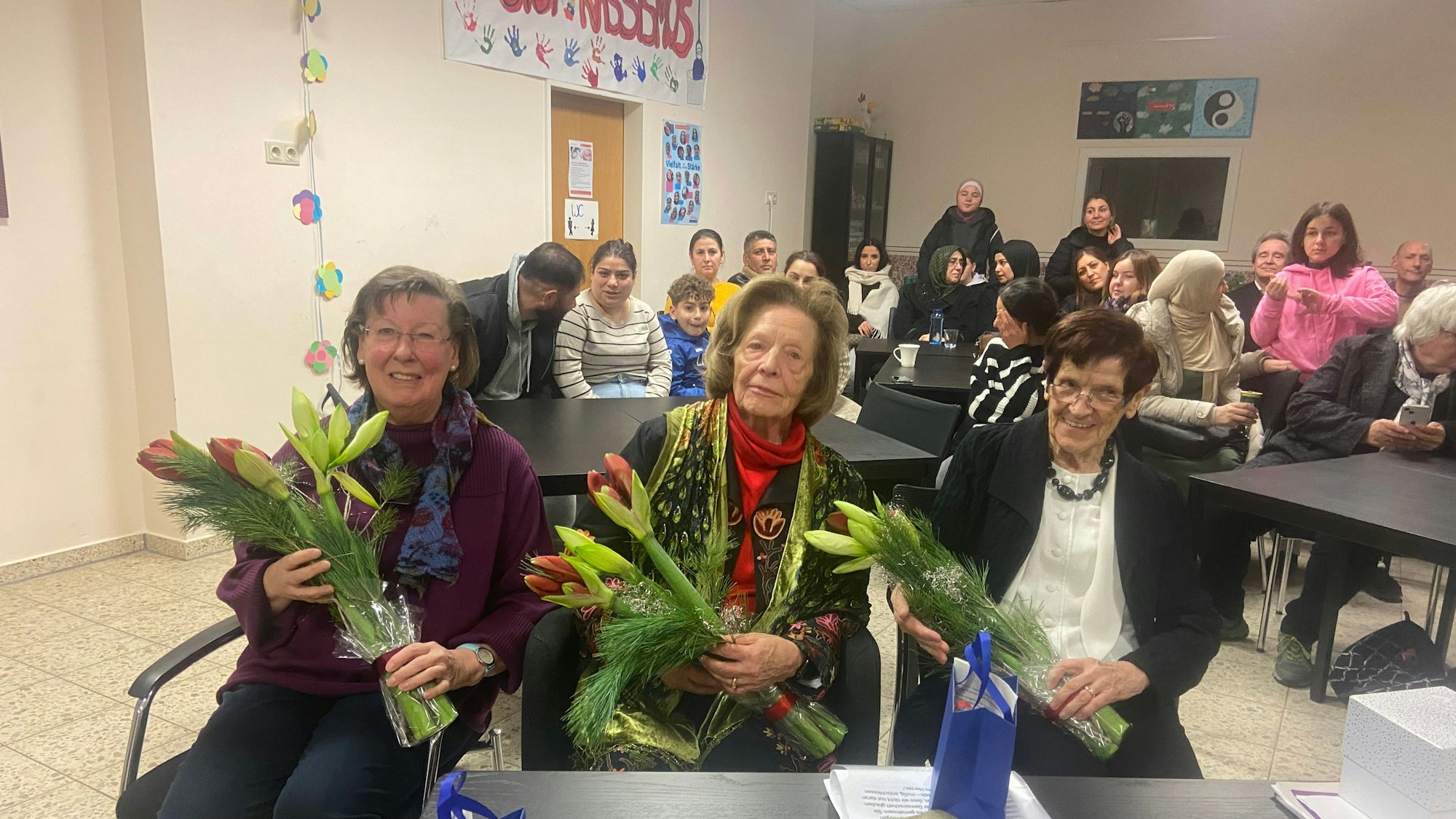 Drei ältere Frauen sitzen vor einem Tisch und halten Blumensträuße.