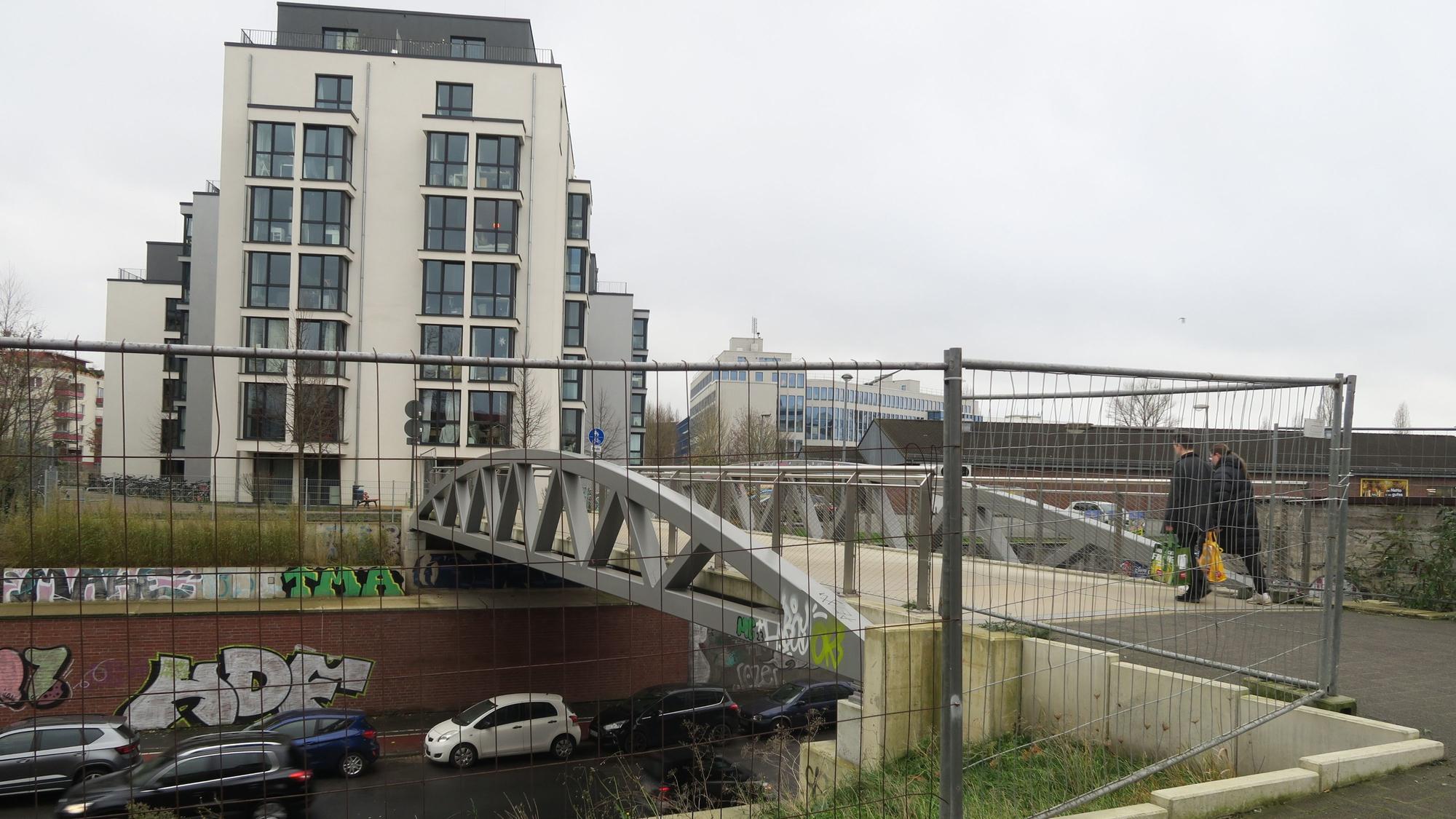 Blick auf eine Fußgängerbrücke, darunter fahren Autos, im Vordergrund steht ein Bauzaun.