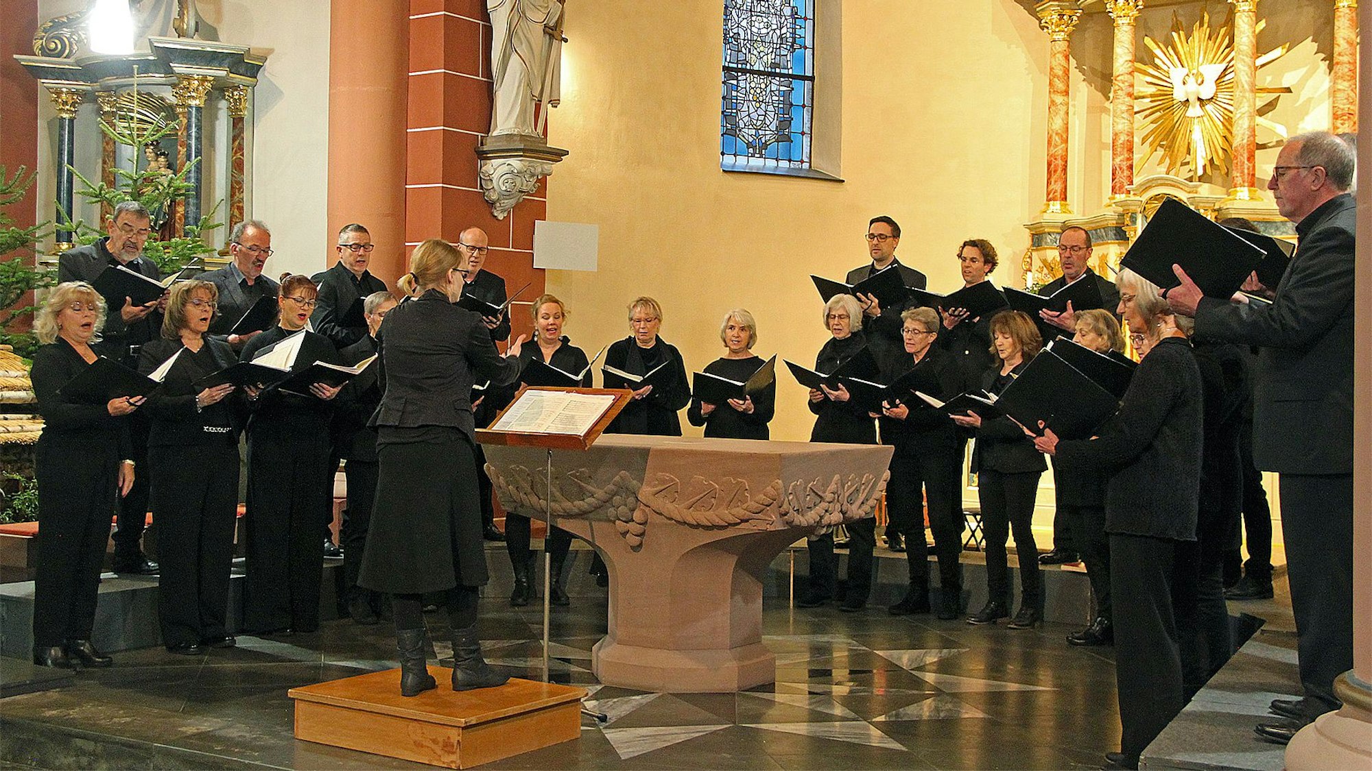Die schwarz gekleideten Mitglieder des gemischten Chores stehen singend im Altarraum einer Kirche, in den Händen halten sie Notenblätter. Die Dirigentin steht auf einem Podest vor dem Altar.