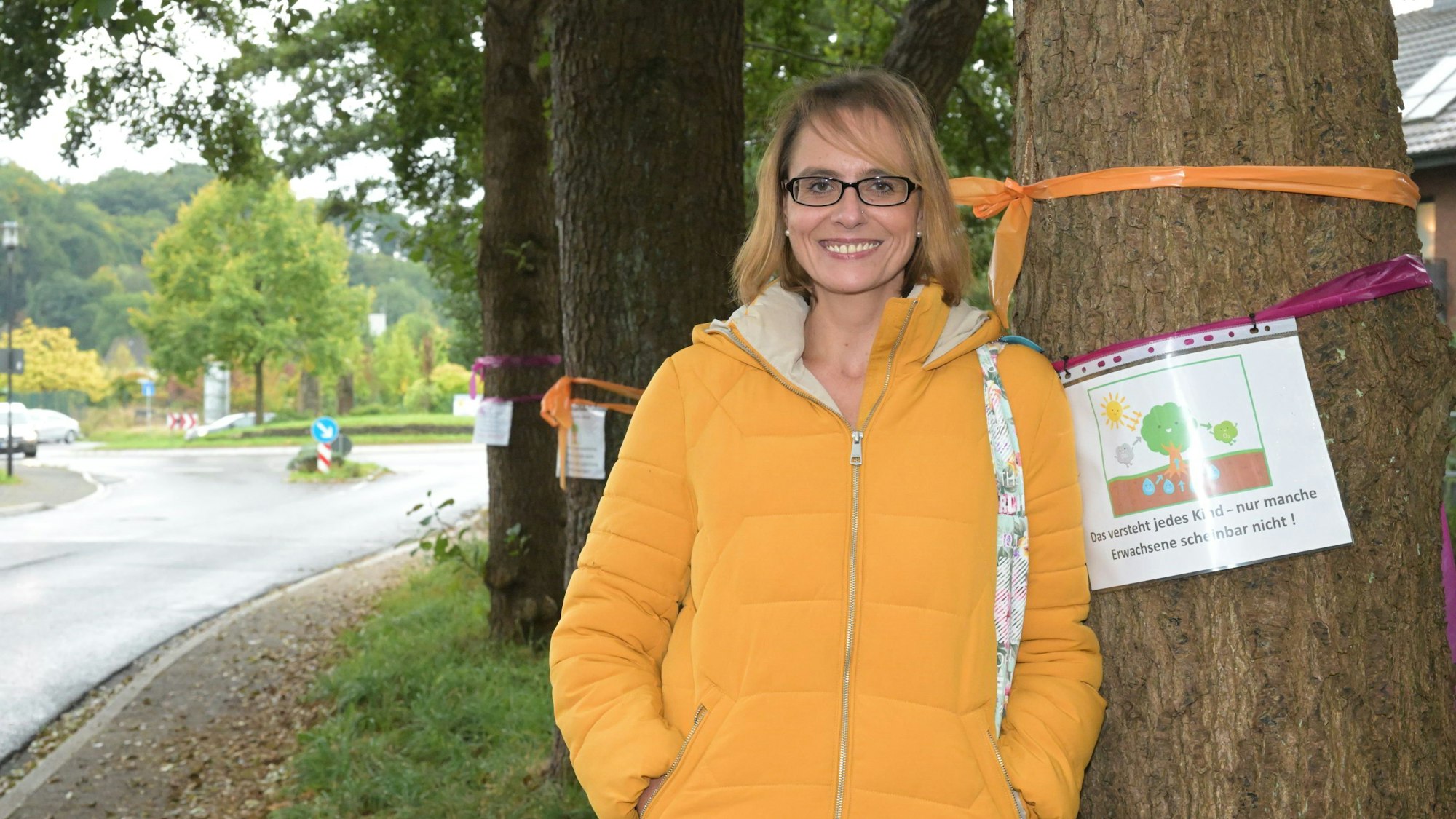Eine Frau in gelber Jacke steht vor alten Bäumen. An den Stämmen hängen Plakate gegen die Fällung der Bäume