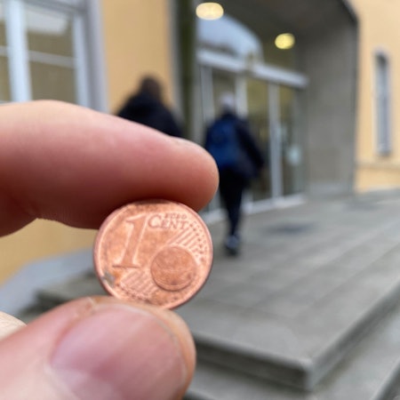 Zu sehen ist eine Ein-Cent-Münze und im Hintergrund das Brühler Rathaus.