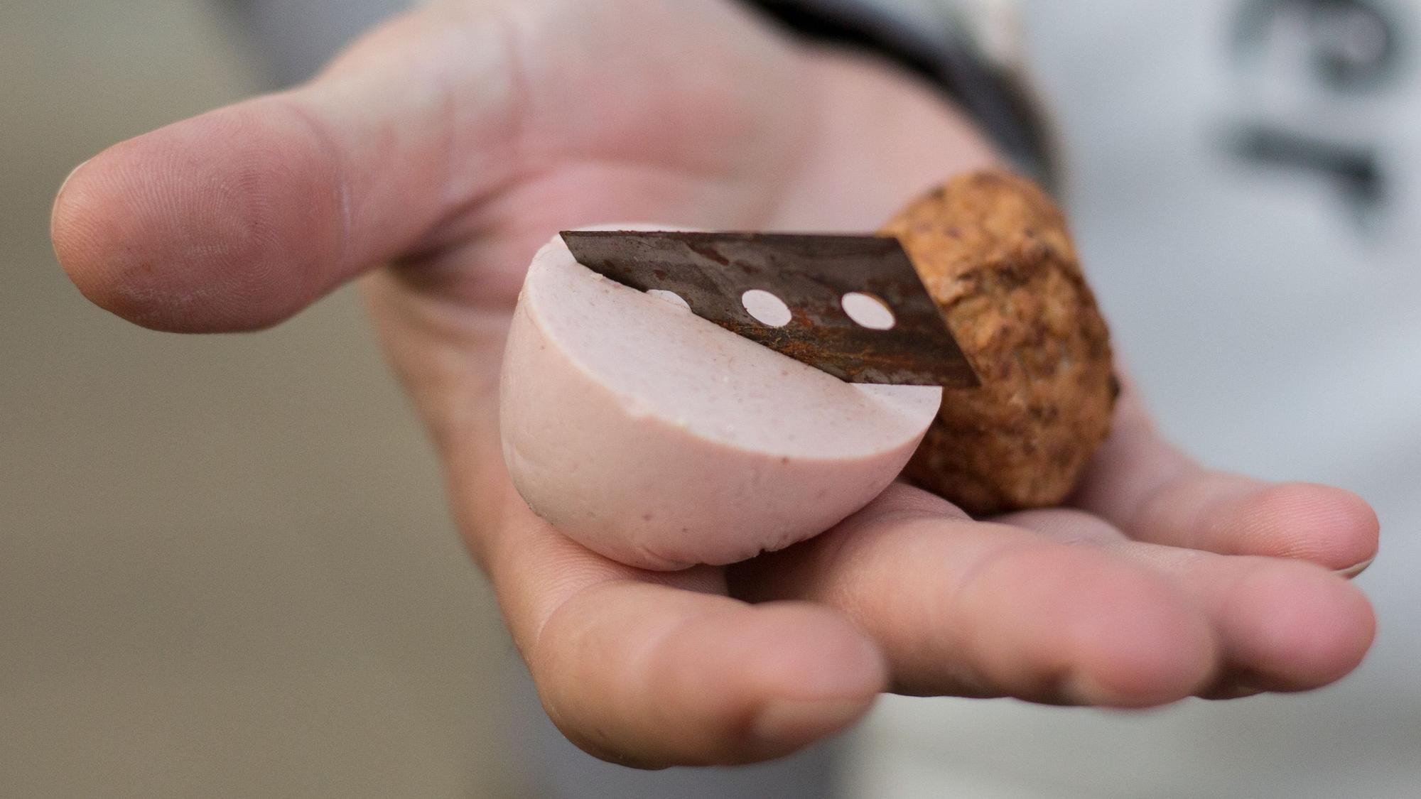 Ein Mann hält ein mit einer Rasierklinge präpariertes Stück Fleischwurst in der Hand.