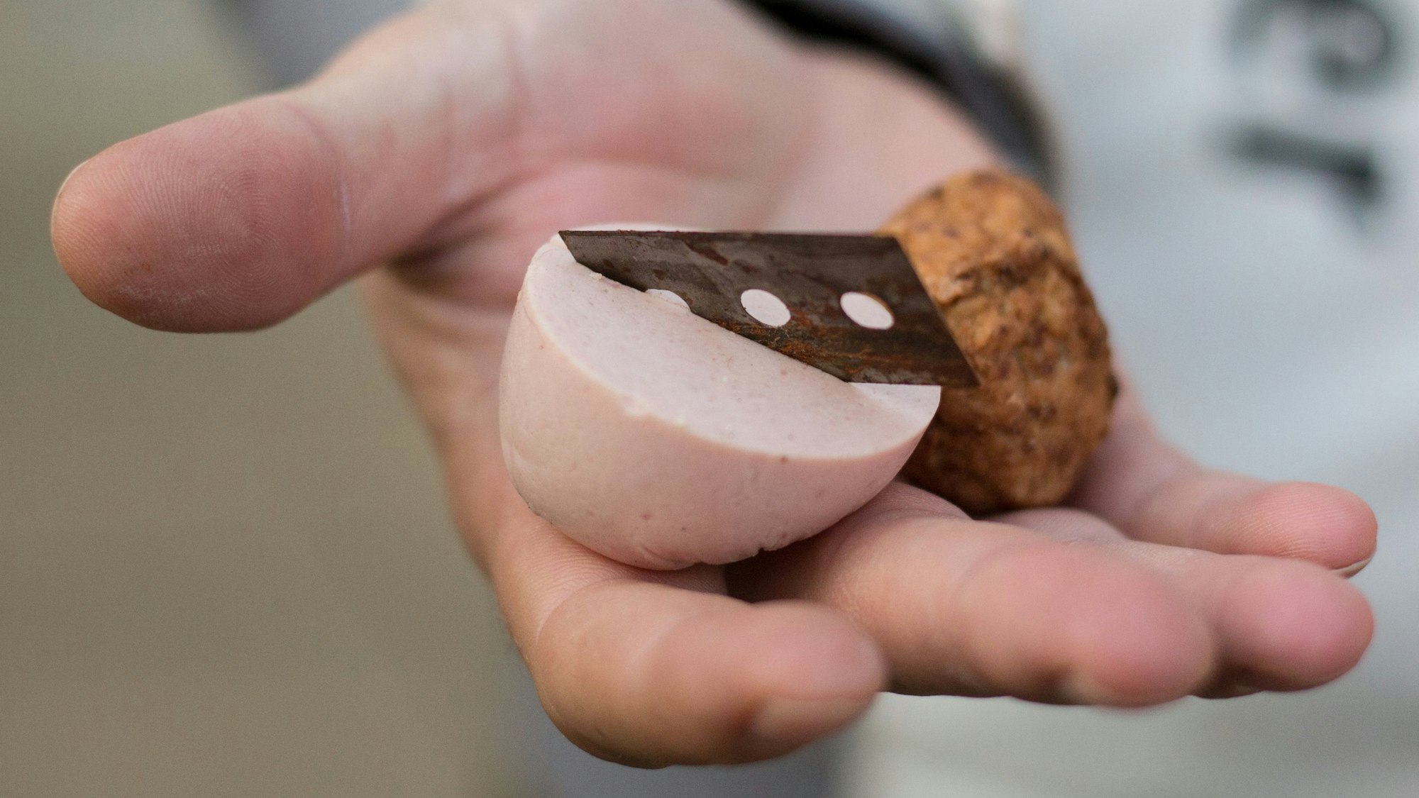Ein Mann hält ein mit einer Rasierklinge präpariertes Stück Fleischwurst in der Hand.