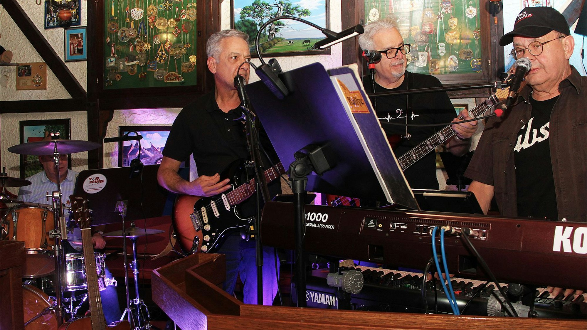 Eine Band aus vier älteren Herren steht eng beieinander in einer Kneipe und macht Musik.