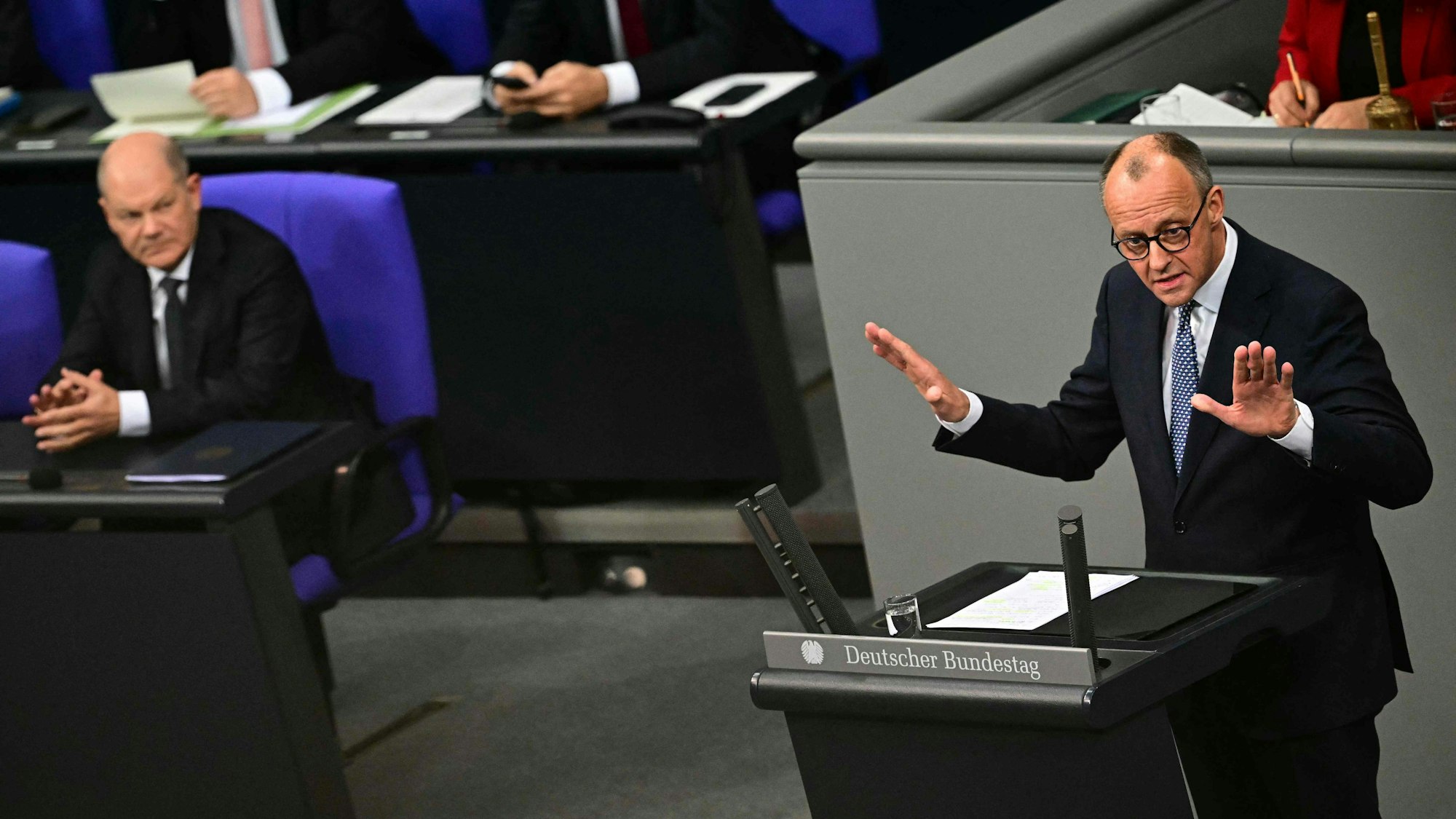 Friedrich Merz hält eine Rede im Bundestag, während Olaf Scholz ihm von der Regierungsbank aus zuschaut.