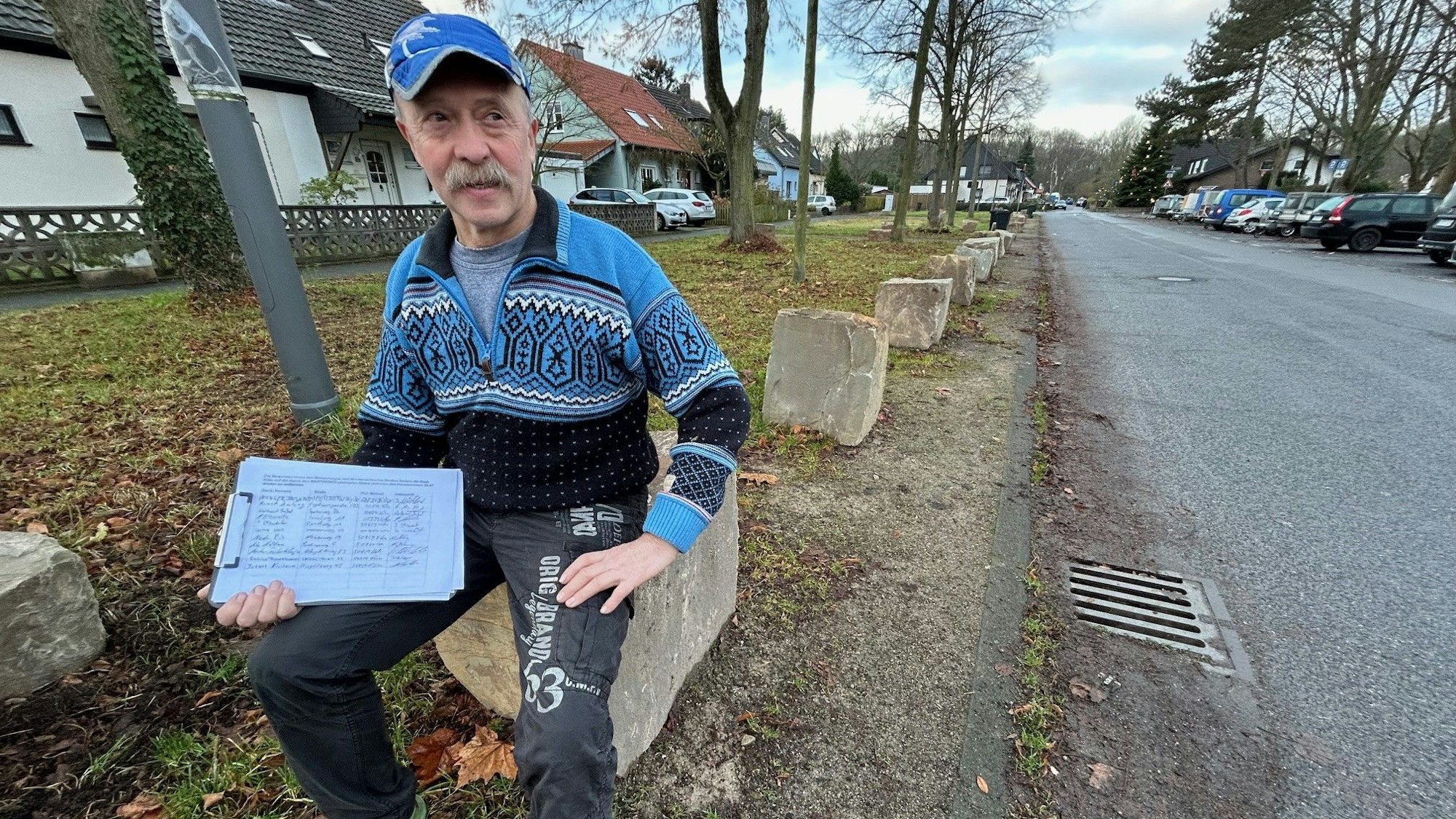Anwohner Gerd Mausbach sitzt mit der Unterschriftenliste auf einem der Felsbrocken, die das Parken auf dem Randstreifen am Meisenweg in Vogelsang unmöglich machen.