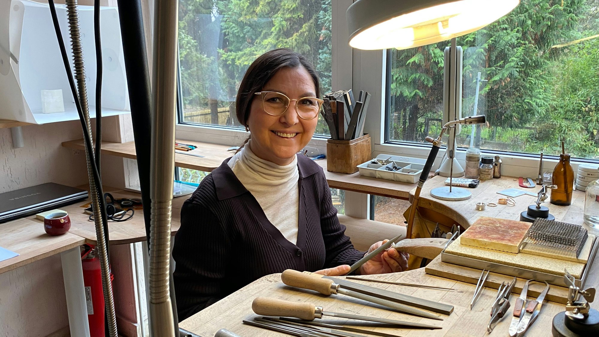 Eine Frau - sie ist Goldschmiedemeisterin - sitzt in ihrer Werkstatt in Frechen, sie lächelt in die Kamera.