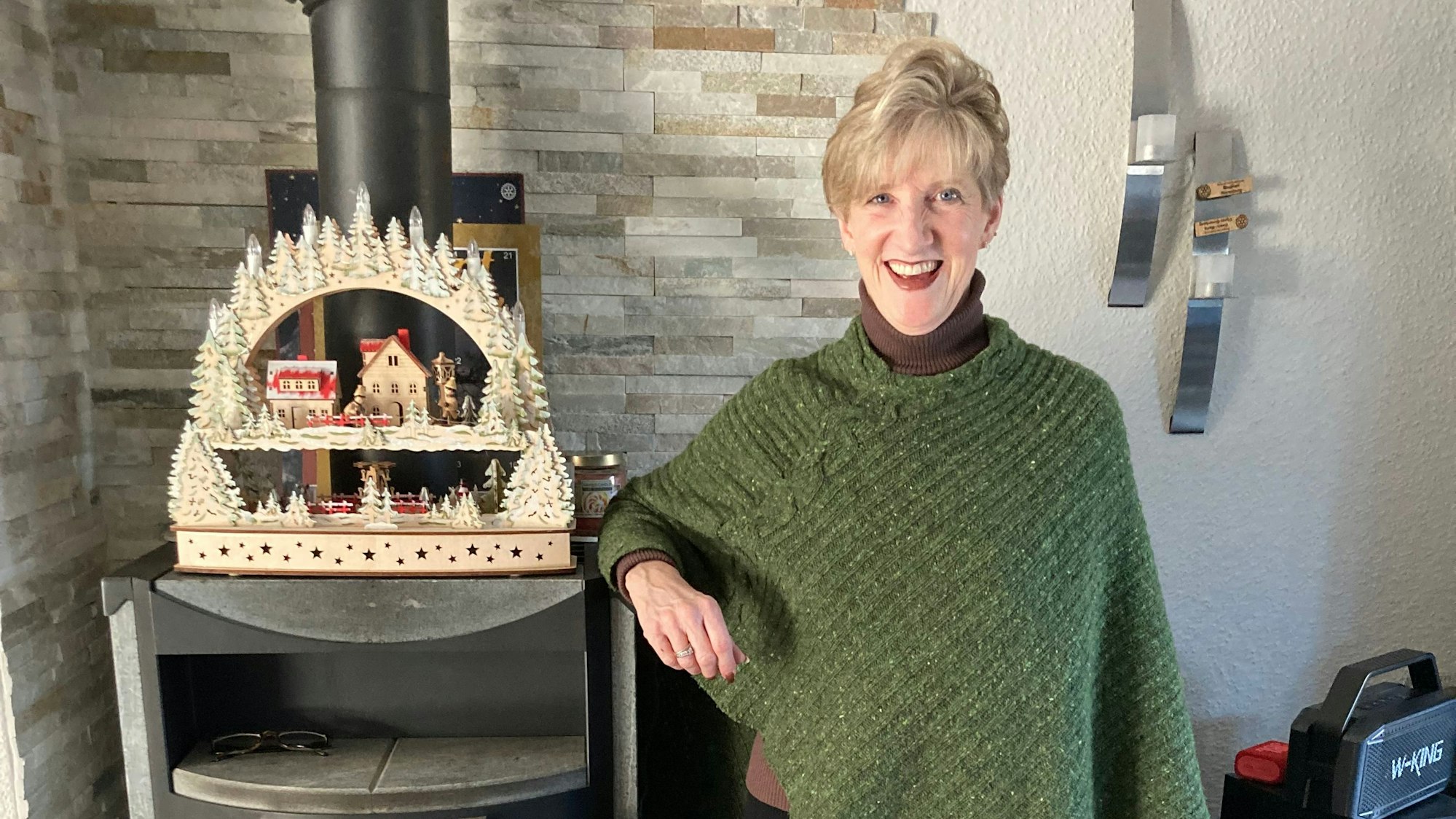 Die blonde Frau steht neben einem Ofen, auf dem ein winterliches Holzbild steht. Sie trägt einen braunen Rollkragenpullover und darüber einen grünen, gestrickten Poncho.