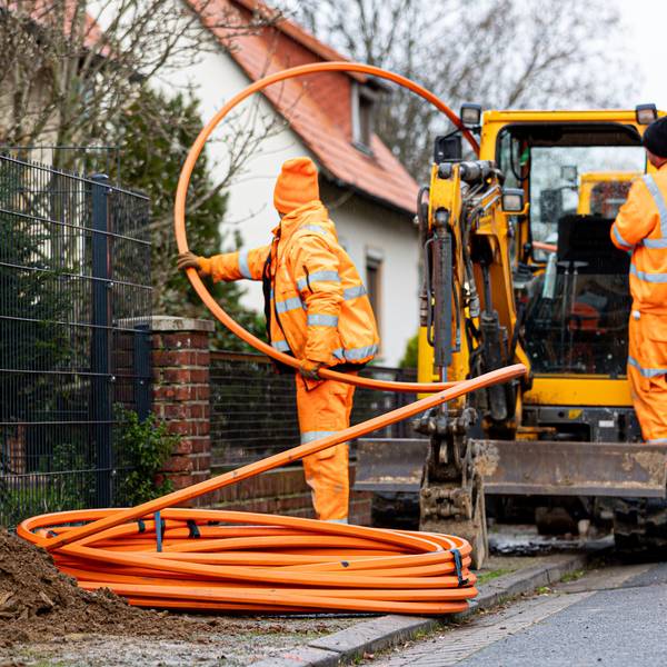 Arbeiter sind mit Bagger und Spaten an einer Straße mit der Erdverlegung von Glasfaserkabeln beschäftigt.