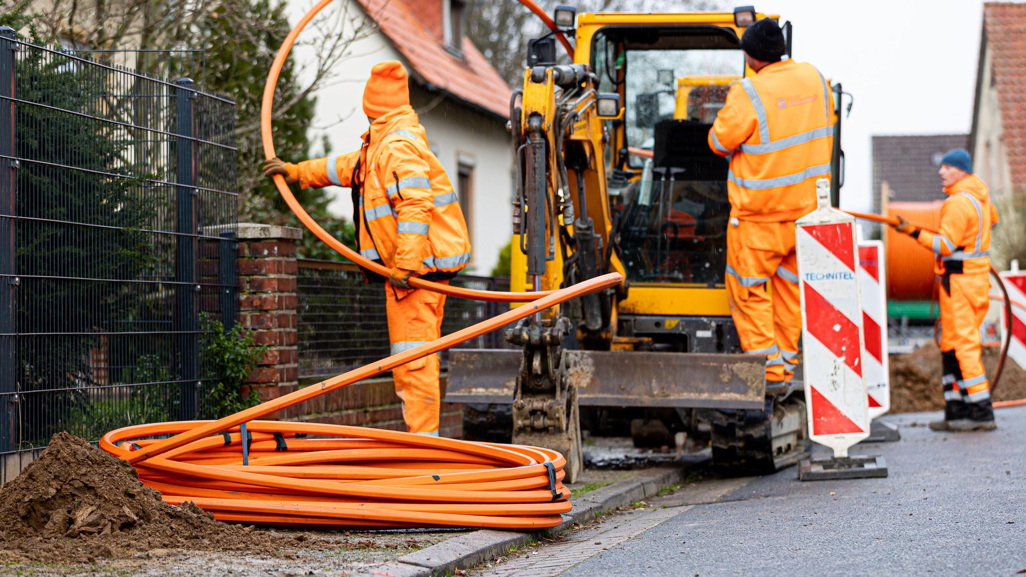 Arbeiter sind mit Bagger und Spaten an einer Straße mit der Erdverlegung von Glasfaserkabeln beschäftigt.
