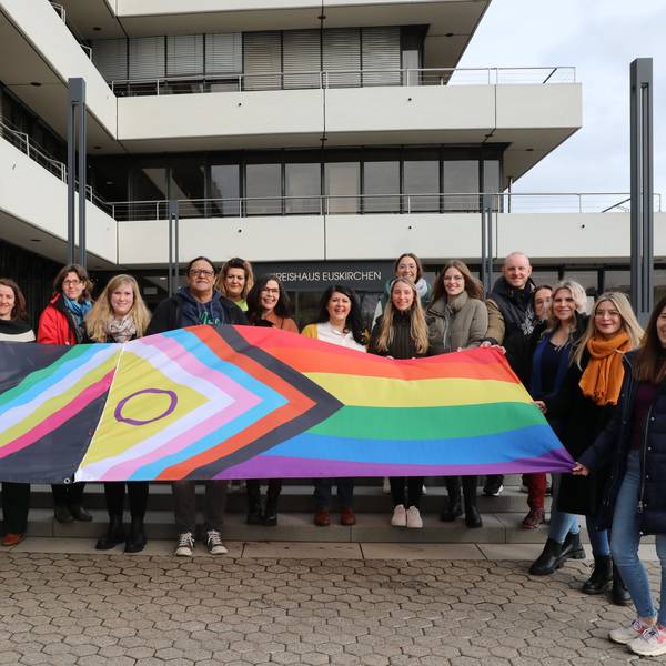 Vor der Treppe des Kreishauses steht eine 19-köpfige Gruppe aus Frauen und Männern. Sie halten eine ausgebreitete Flagge, die mit ihren Farben und Symbolen für die Vielfalt der Menschen steht.