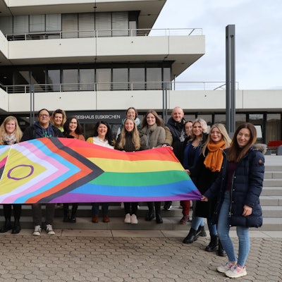 Vor der Treppe des Kreishauses steht eine 19-köpfige Gruppe aus Frauen und Männern. Sie halten eine ausgebreitete Flagge, die mit ihren Farben und Symbolen für die Vielfalt der Menschen steht.