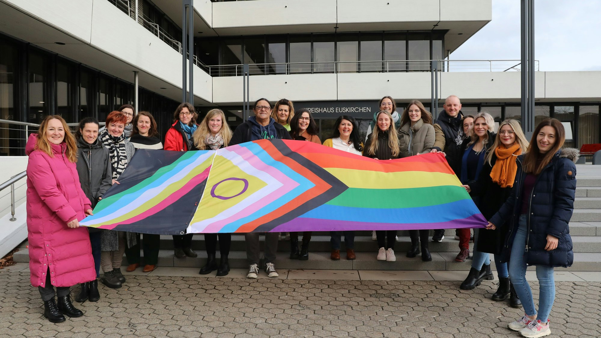 Vor der Treppe des Kreishauses steht eine 19-köpfige Gruppe aus Frauen und Männern. Sie halten eine ausgebreitete Flagge, die mit ihren Farben und Symbolen für die Vielfalt der Menschen steht.