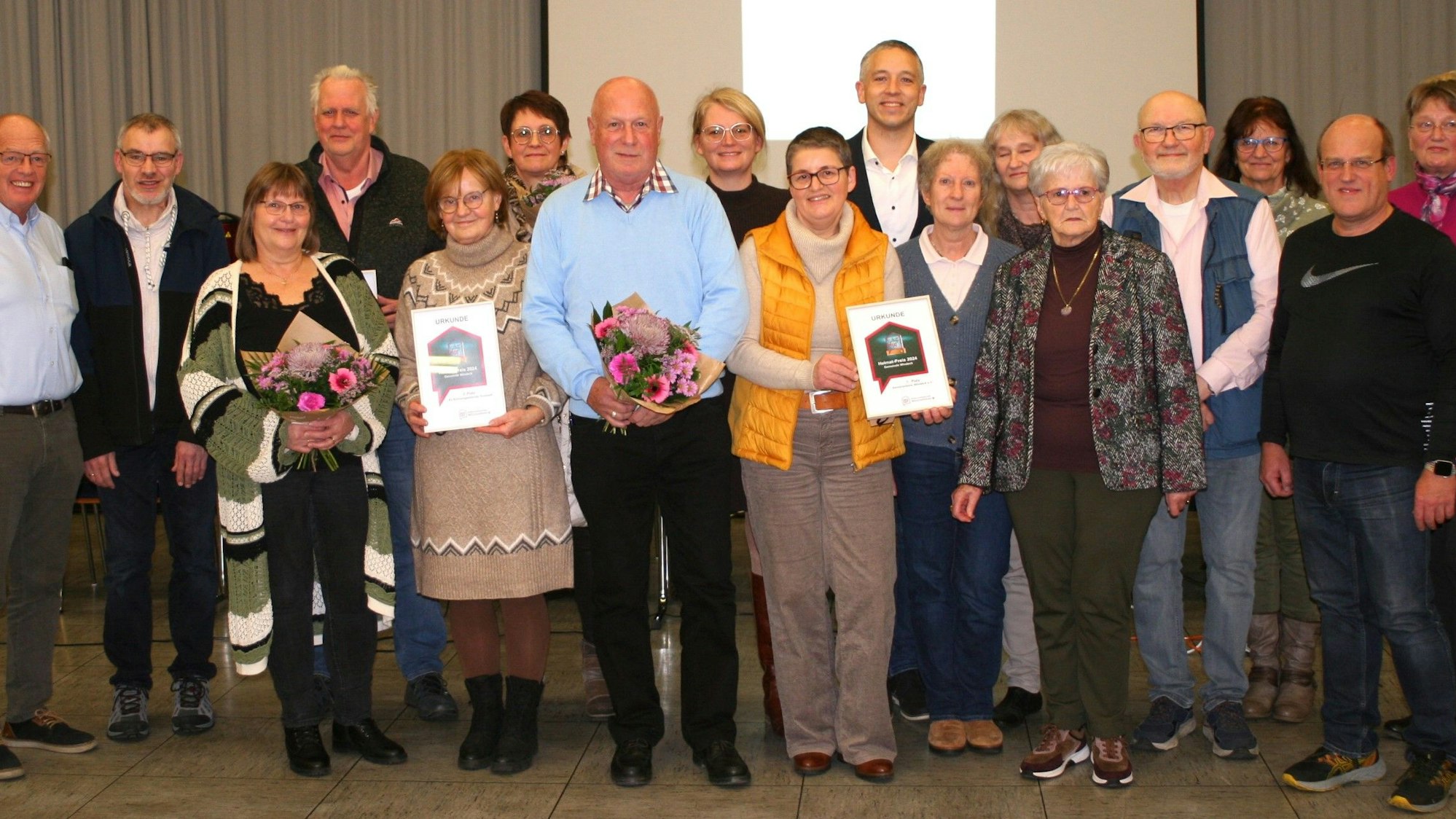 17 Männer und Frauen stehen für ein Gruppenfoto zusammen, in der vorderen Reihe werden Blumensträuße und Urkunden gehalten.