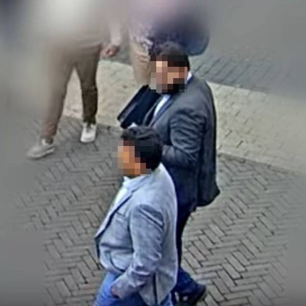 Auf der Aufnahme einer Überwachungskamera sind zwei Männer zu sehen, die Anzüge tragen. Die Gesichter der Männer sind verpixelt.