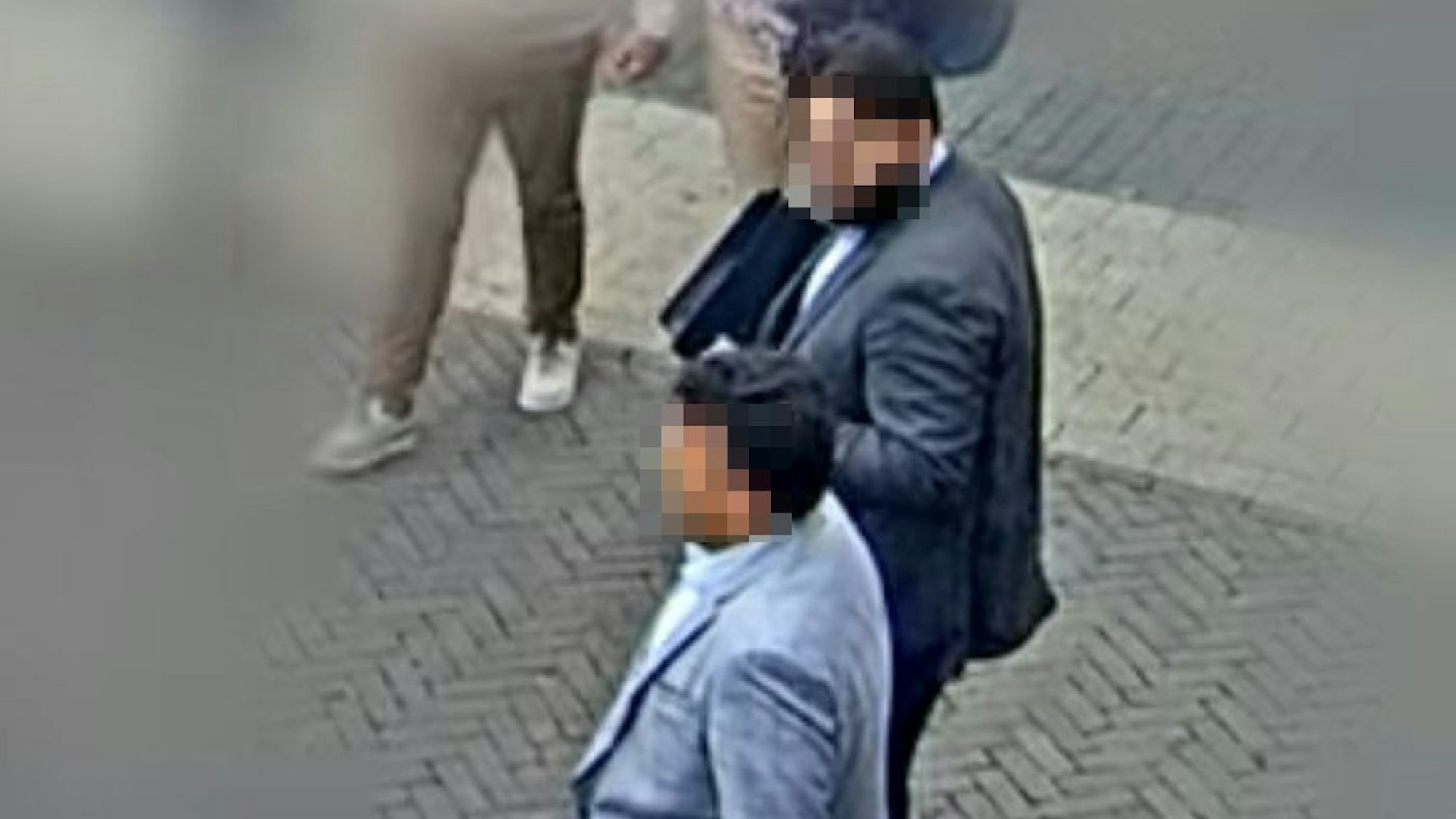 Auf der Aufnahme einer Überwachungskamera sind zwei Männer zu sehen, die Anzüge tragen. Die Gesichter der Männer sind verpixelt.