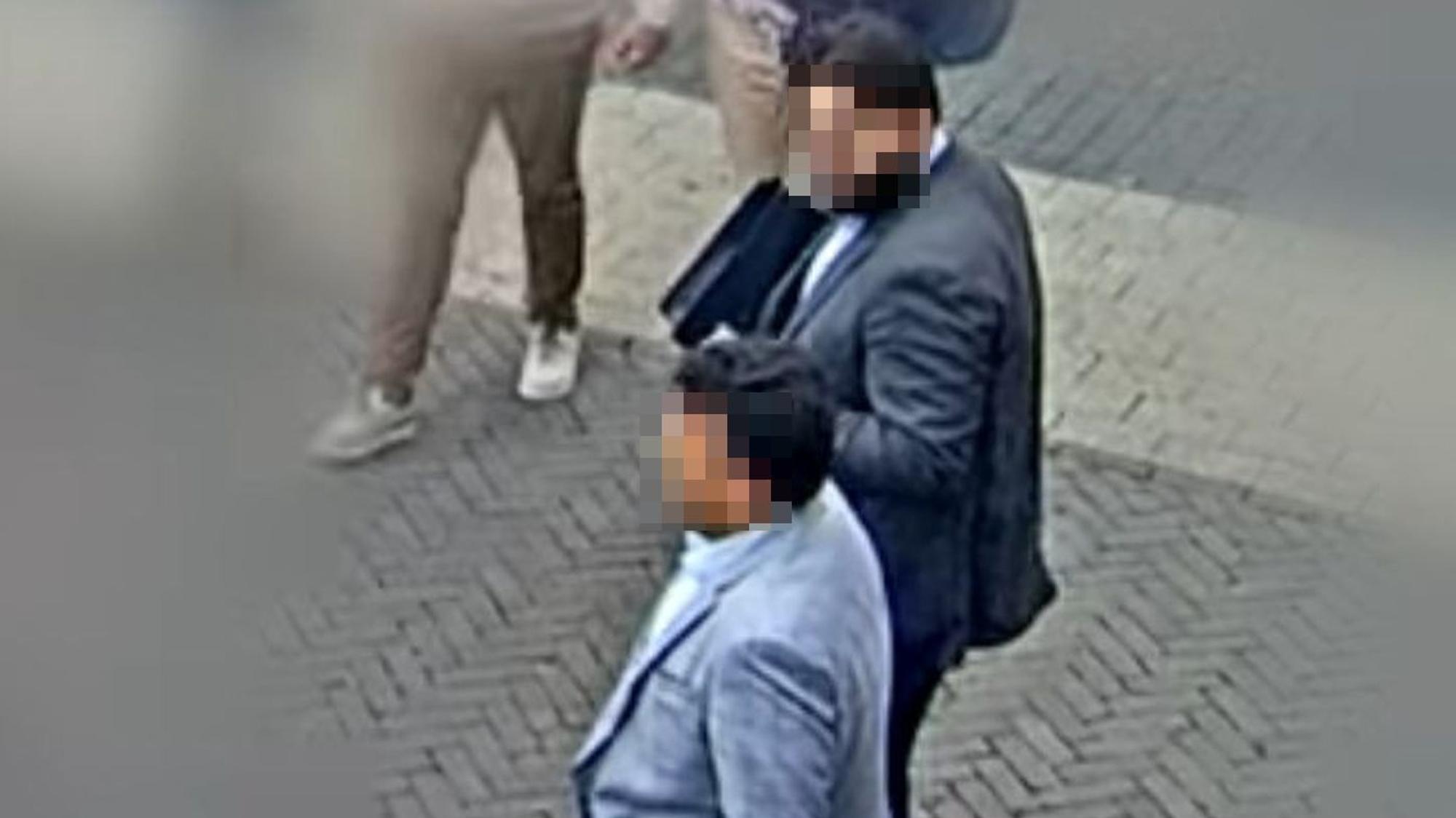 Das Bild einer Überwachungskamera zeigt zwei Männer unterschiedlicher Größe, die Anzüge tragen. Die Gesichter sind verpixelt dargestellt.
