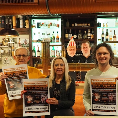 Sechs Männer und eine Frau haben sich vor und hinter der Theke verteilt. Einige von ihnen präsentieren das Veranstaltungsplakat.