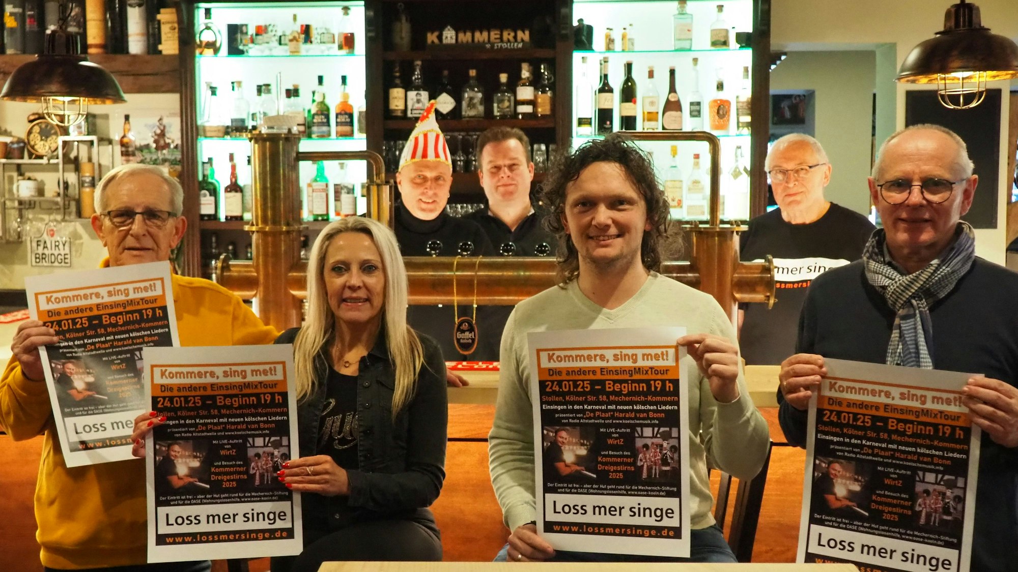 Sechs Männer und eine Frau haben sich vor und hinter der Theke verteilt. Einige von ihnen präsentieren das Veranstaltungsplakat.