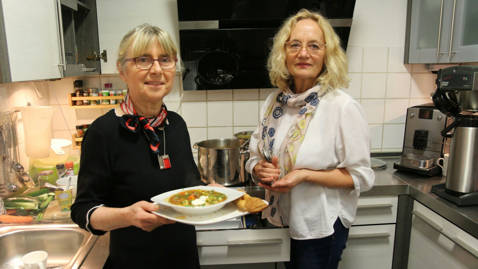 Zwei Frauen stehen in einer Küche, eine hält einen Teller mit Suppe in den Händen.