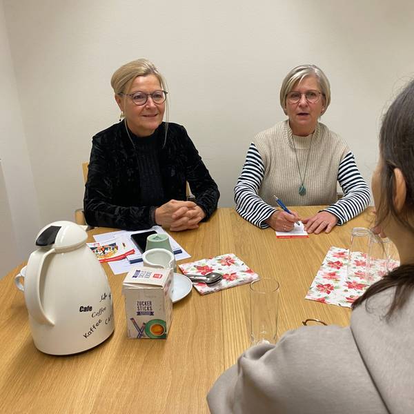 Drei Frauen sitzen an einem Tisch, eine mit dem Rücken zum Betrachter. Auf dem Tisch steht eine Thermoskanne.