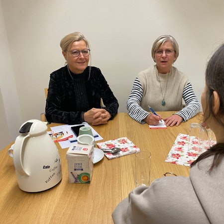 Drei Frauen sitzen an einem Tisch, eine mit dem Rücken zum Betrachter. Auf dem Tisch steht eine Thermoskanne.