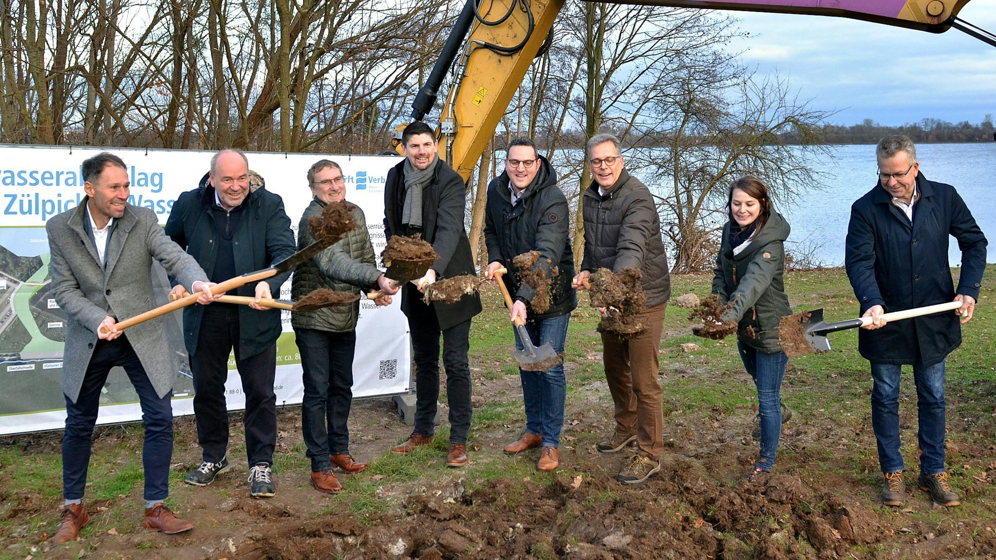 Eine Gruppe Männer und eine Frau stehen auf einer Wiese und halten Schaufeln mit Erde darauf in der Hand. Im Hintergrund ist die Oberfläche eines Sees zu erkennen.