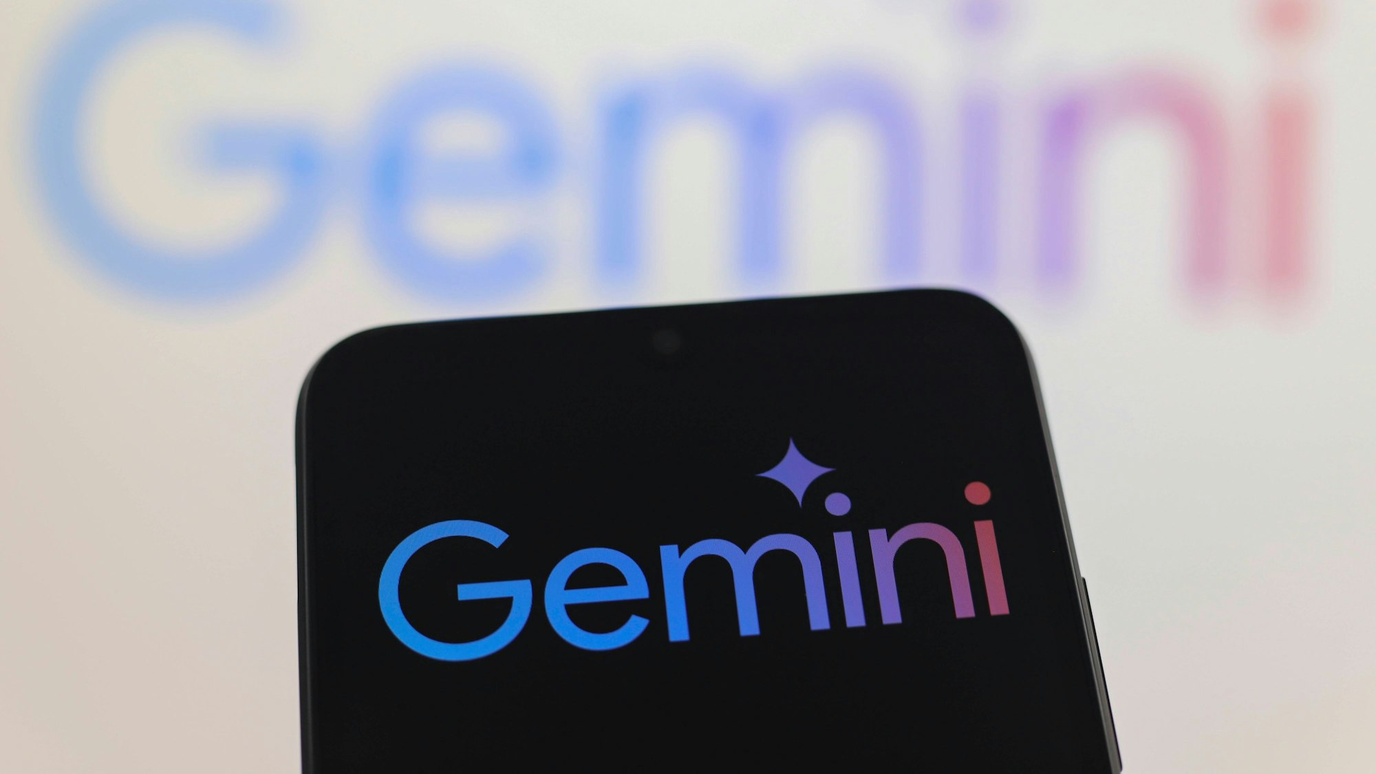 Ein Smartphone mit dem Logo von Gemini vor einer Wand, auf der ebenfalls Gemini steht.