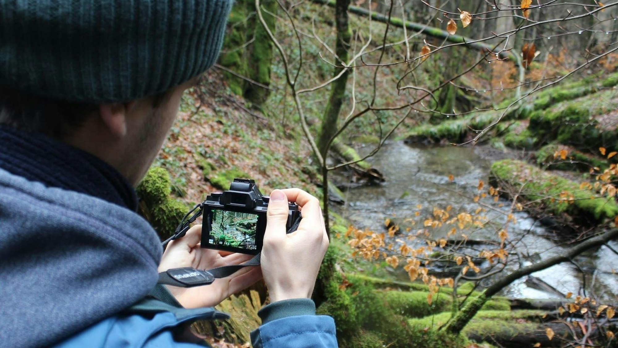 An einem Bach im Nationalpark Eifel steht ein Mann, der ein Foto mit einer Digitalkamera aufnimmt.