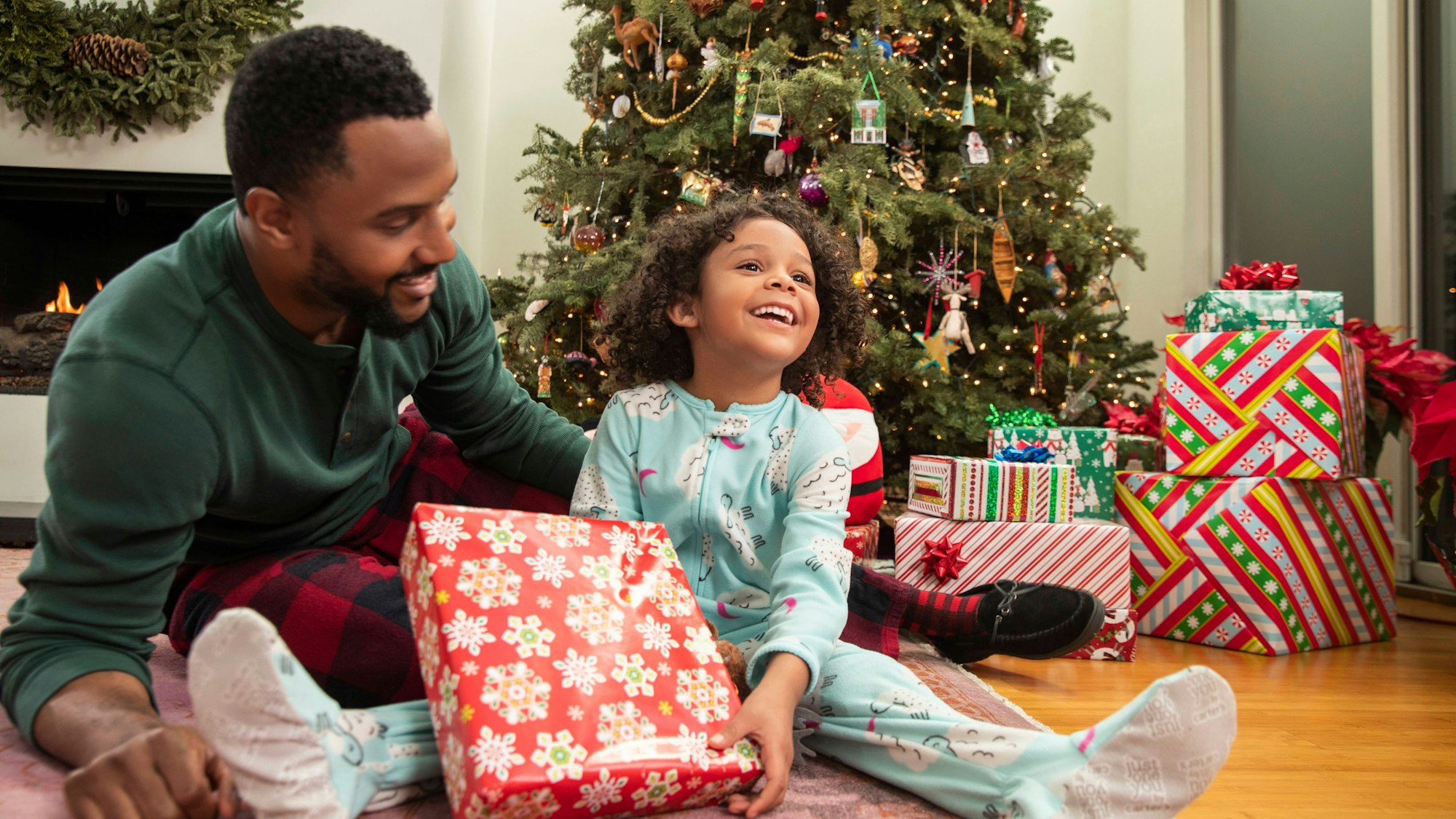 Ein Vater und seine junge Tochter sitzen vor einem Weihnachtsaum, beide strahlen, auf dem Bild sind viele Geschenke zu sehen.