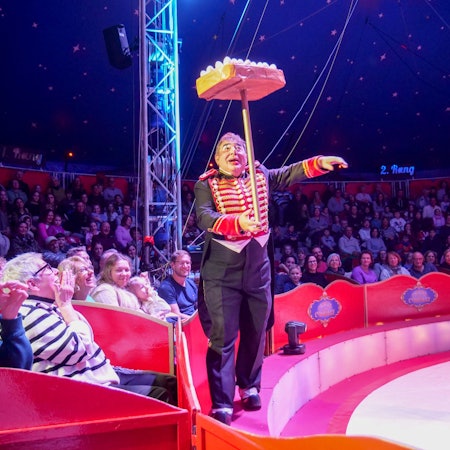 Das Foto zeigt den Clown mit einer Jonglage-Nummer vor den Rängen rund um die Manege.