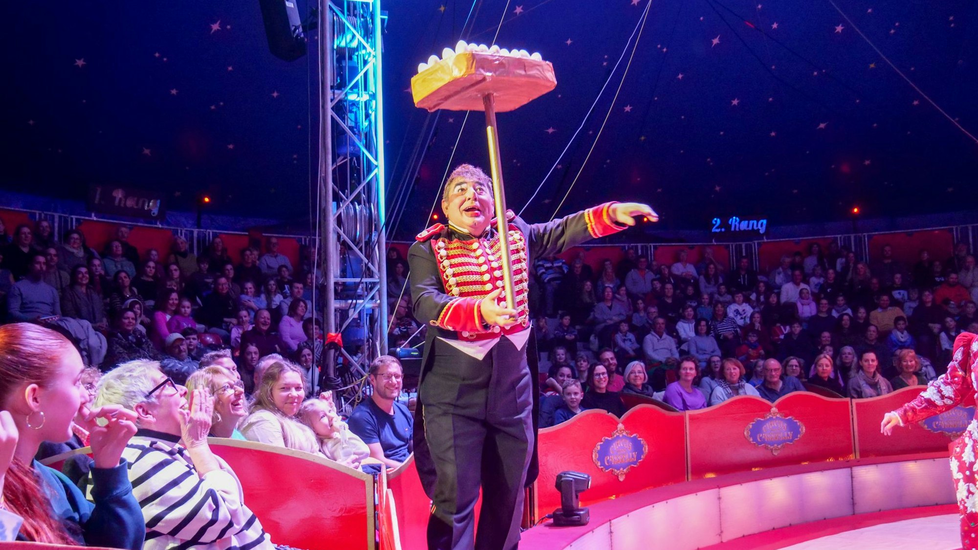 Das Foto zeigt den Clown mit einer Jonglage-Nummer vor den Rängen rund um die Manege.