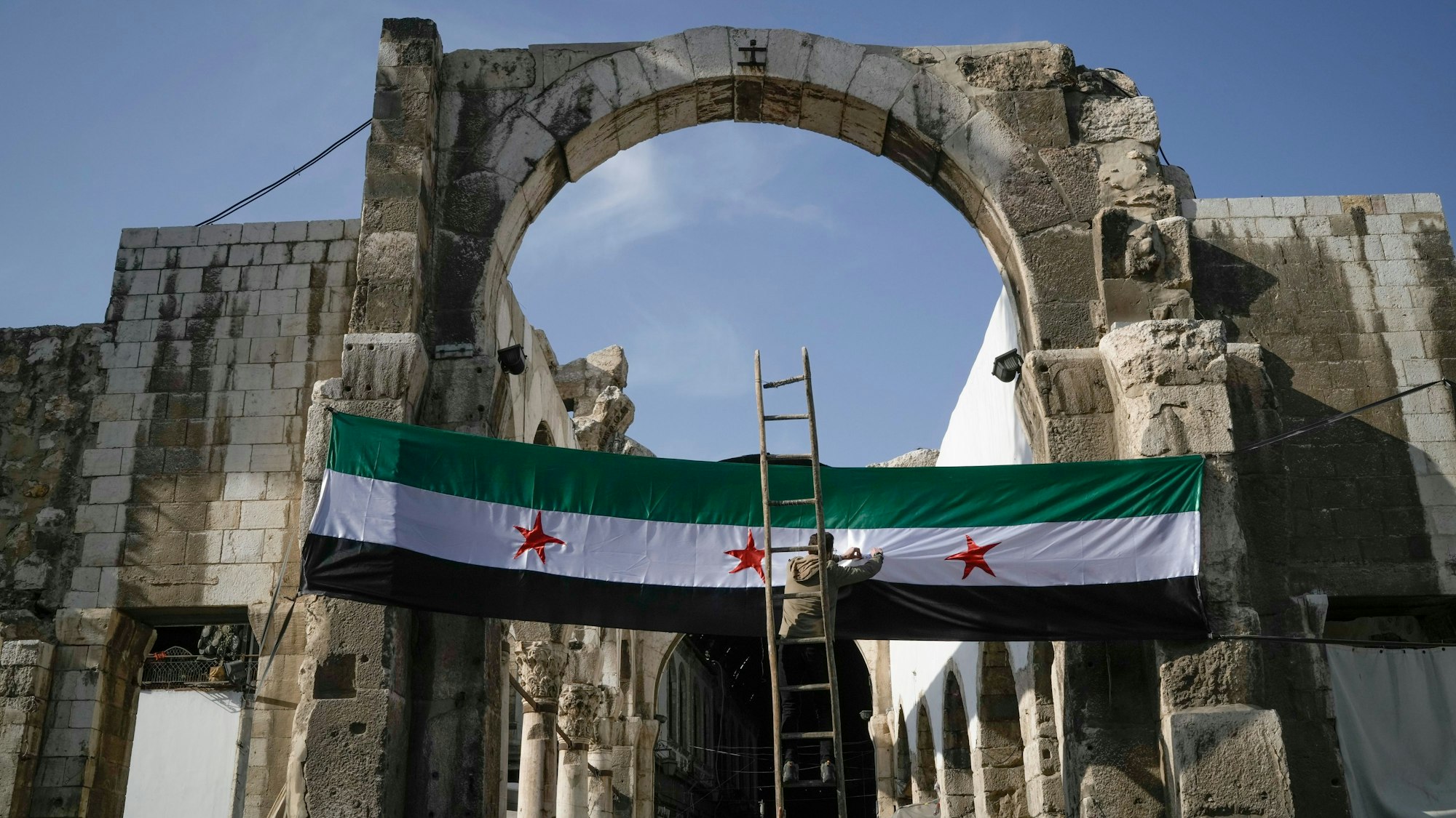 20.12.2024, Syrien, Damaskus: Ein Arbeiter hängt eine Flagge der syrischen Revolutionäre - grün, weiß, schwarz mit roten Sternen am Eingang zu dem Suq al-Hamidiya, vor dem Freitagsgebet auf. Foto: Leo Correa/AP/dpa +++ dpa-Bildfunk +++
