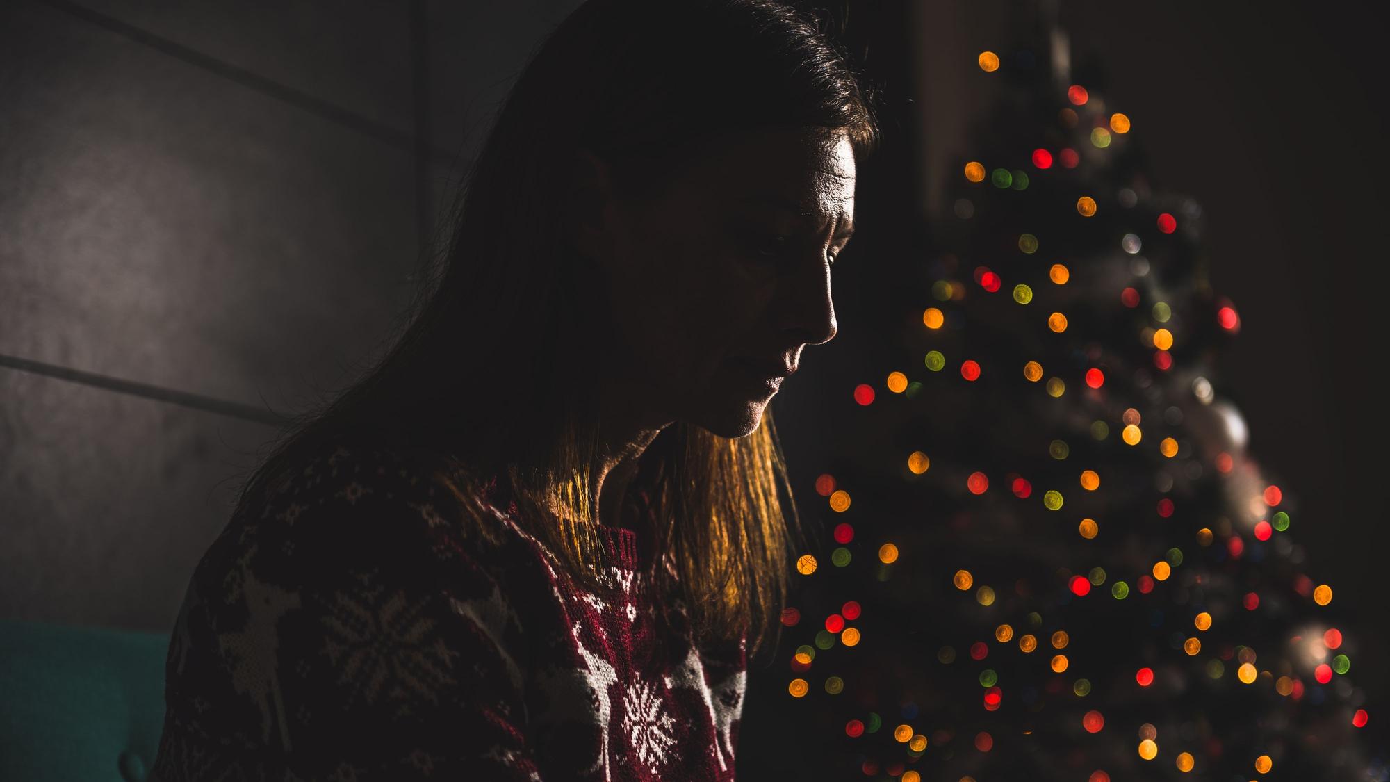Eine Frau sitzt allein in einem dunklen Raum, im Hintergrund ist unscharf ein Weihnachtsbaum zu erkennen.