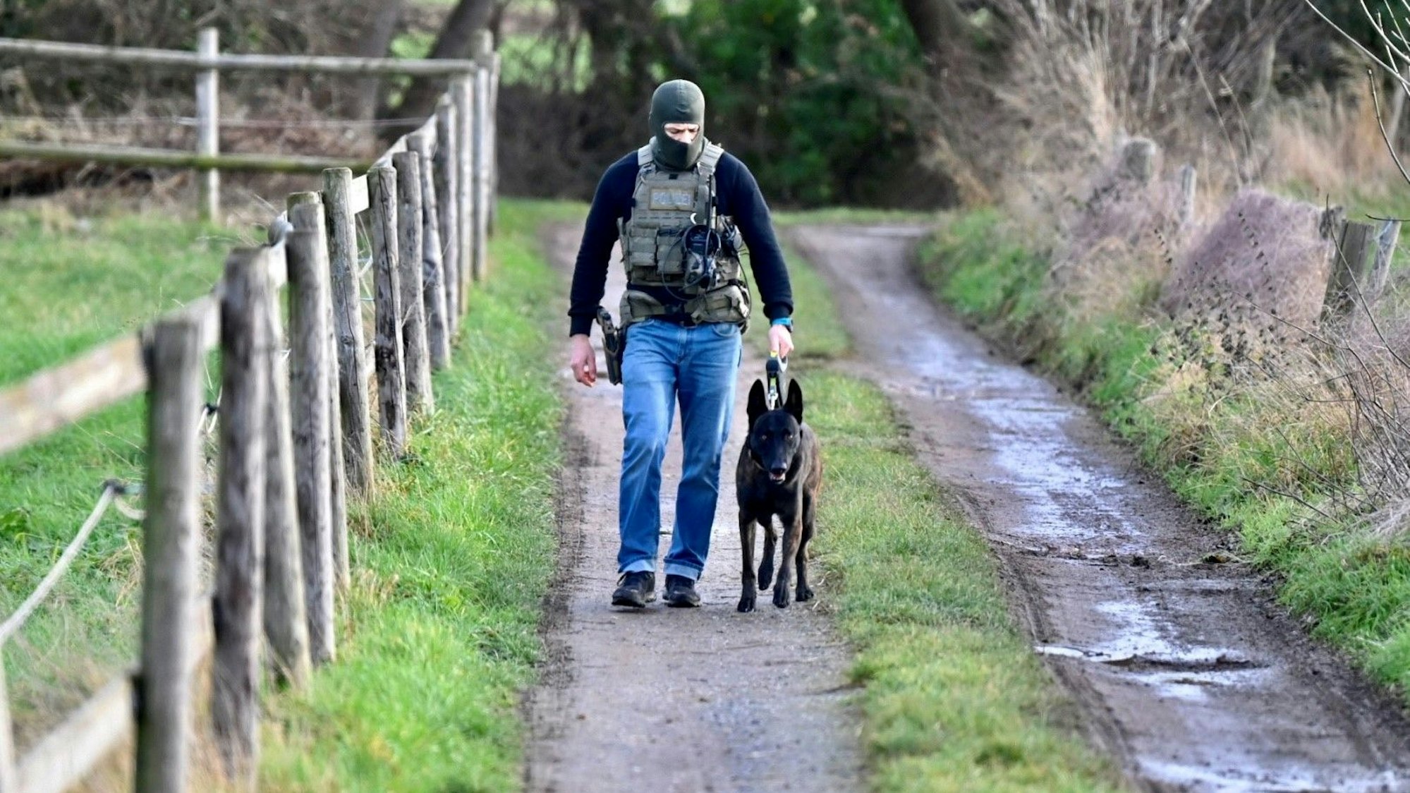 Ein Polizist trägt eine Schutzweste und eine Sturmhaube. Er führt einen Hund an der Leine.