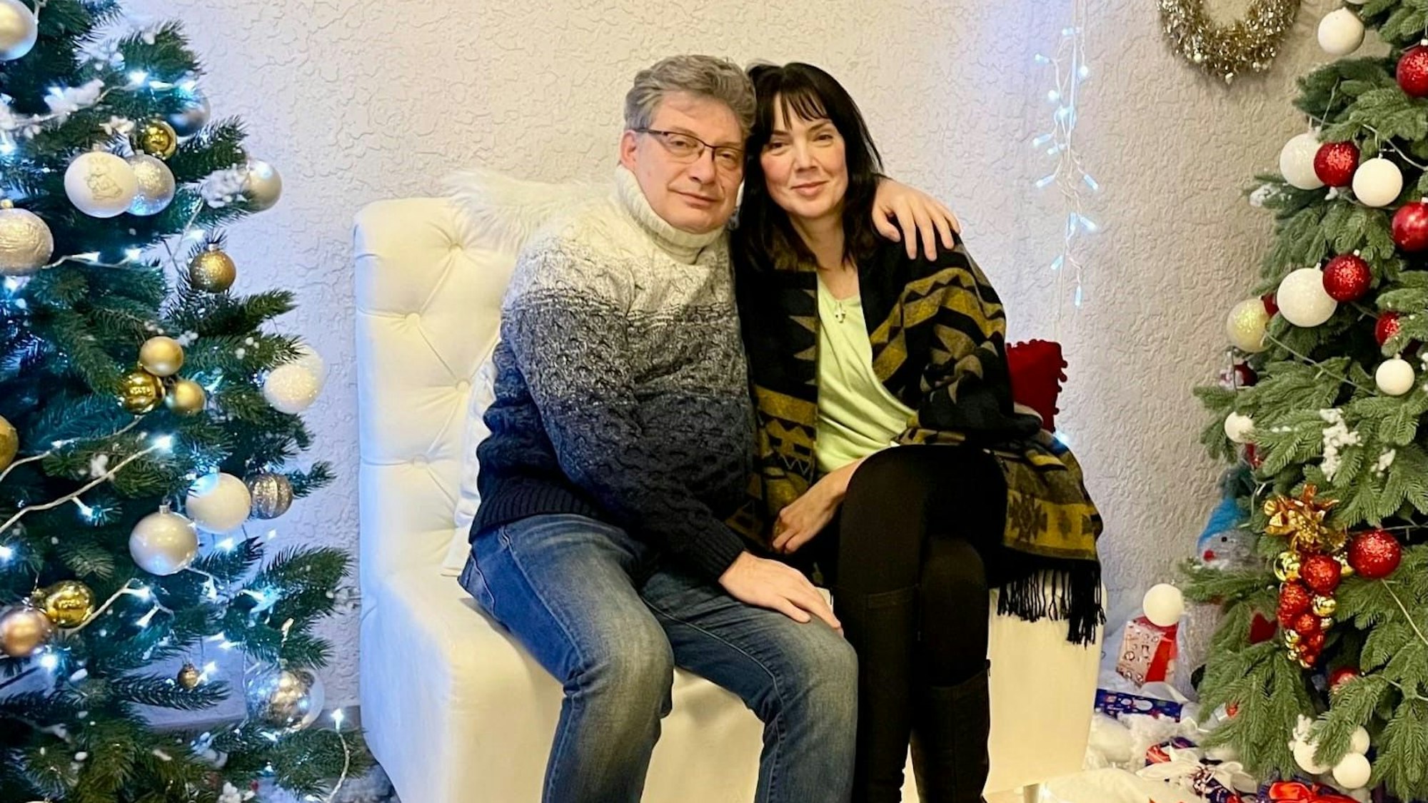 Ein Ehepaar sitz auf einem Sessel. Daneben stehen geschmückte Weihnachtsbäume.