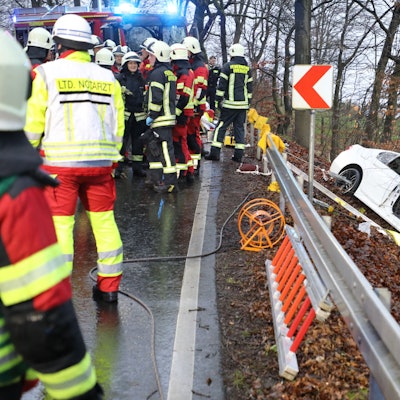 Ein schwer beschädigtes Auto steht vor einem Baum in einer Böschung, auf der Straße oberhalb stehen Feuerwehrleute.