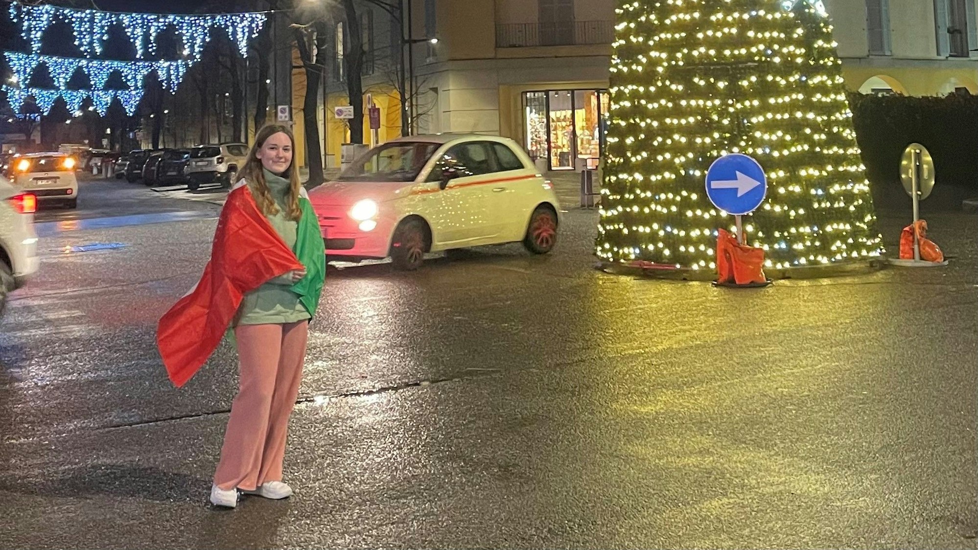 Eine junge Frau mit Italienflagge um die Schultern steht vor einem Weihnachtsbaum inmitten eines Kreisverkehrs.