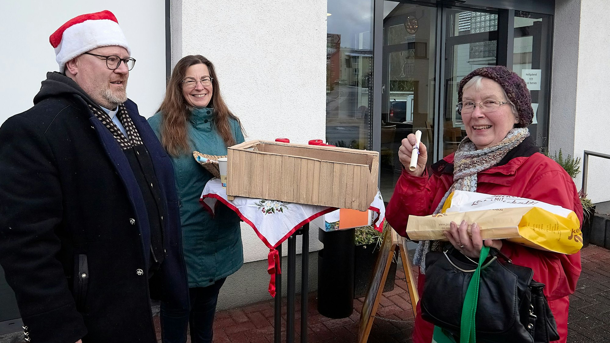 Pfarrer Michael Krosch trägt eine rot-weiße Weihnachtsmütze. Er steht mit Gemeindereferentin Dagmar Goffart und einer weiteren Frau an einem Stehtisch.