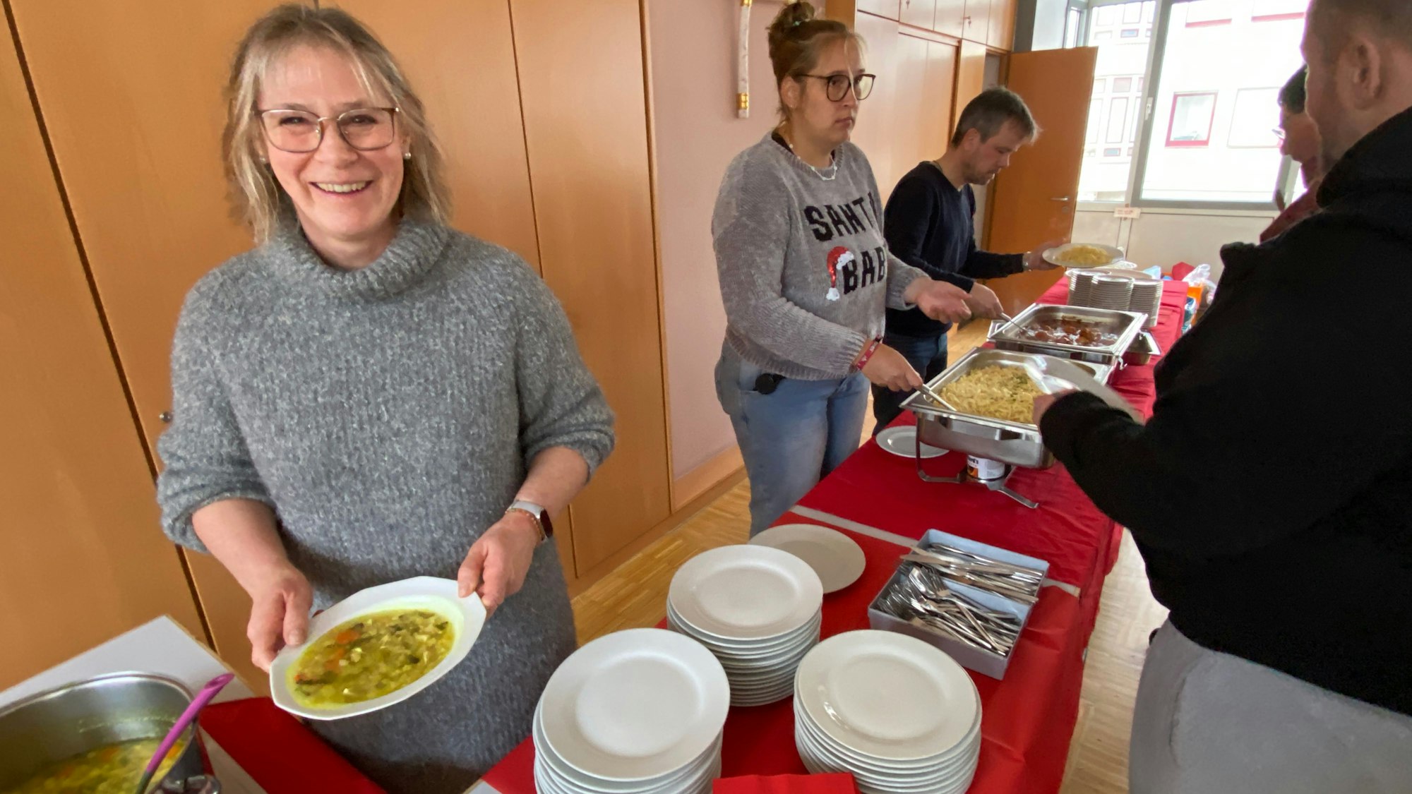 Zwei Frauen und ein Mann stehen hinter einem Tisch mit Tellern und Essen. Sie reichen gefüllte Teller an die davor Wartenden.