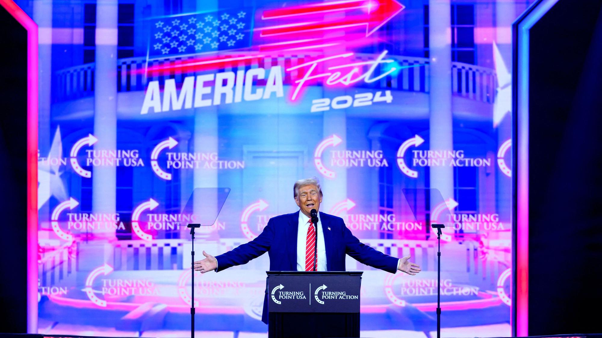 Donald Trump steht auf einer Bühne, im Hintergrund ist eine große LED-Leinwand zu sehen.
