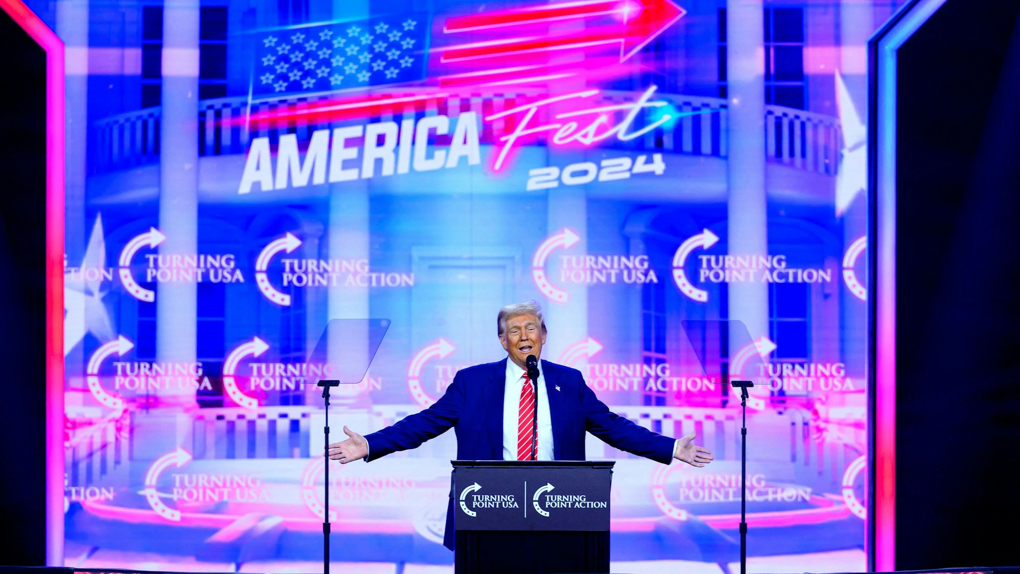 Donald Trump steht auf einer Bühne, im Hintergrund ist eine große LED-Leinwand zu sehen.