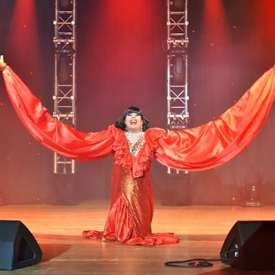 Das Bild zeigt einen Moment in der Show. Eine Künstlerin steht mit überdimensional ausgebreiteten Armen auf der Bühne.